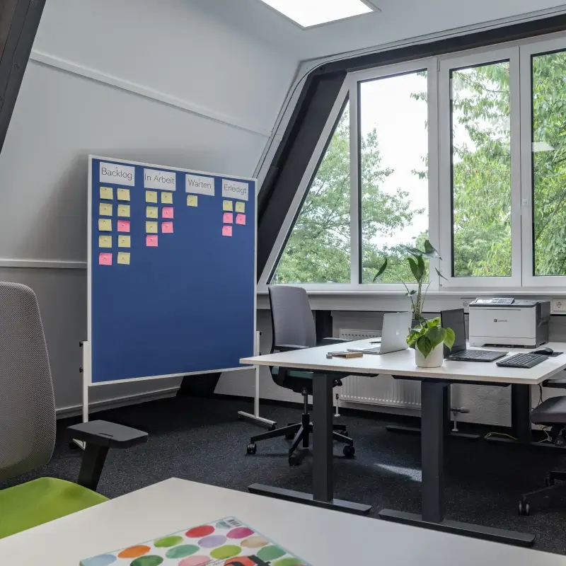 Perspektive von einem dritten Arbeitsplatz im Teambüro mit Schreibtischen, Bürostühlen, Projektmanagement-Pinnwannd, Drucker , Laptops und einer Pflanze.