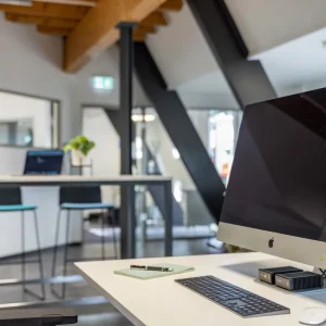 Modern gestalteter Coworking-Schreibtisch im OG2 mit iMac, unterstrichen von natürlichen Holzelementen und schwarzen Stahlträgern, die eine gemütliche, produktive Arbeitsatmosphäre schaffen.