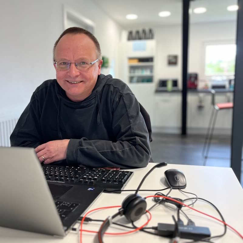 Stephan lächelt, während er an einem Laptop in einem hellen und modernen Coworkingspace arbeitet, umgeben von seinem Headset und weiterem Zubehör.